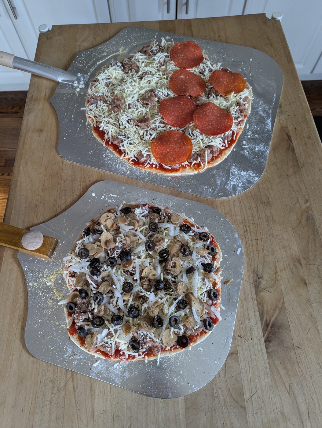 Two pizzas being built side by side on pizza peels, one pepperoni, one with mushrooms and olives
