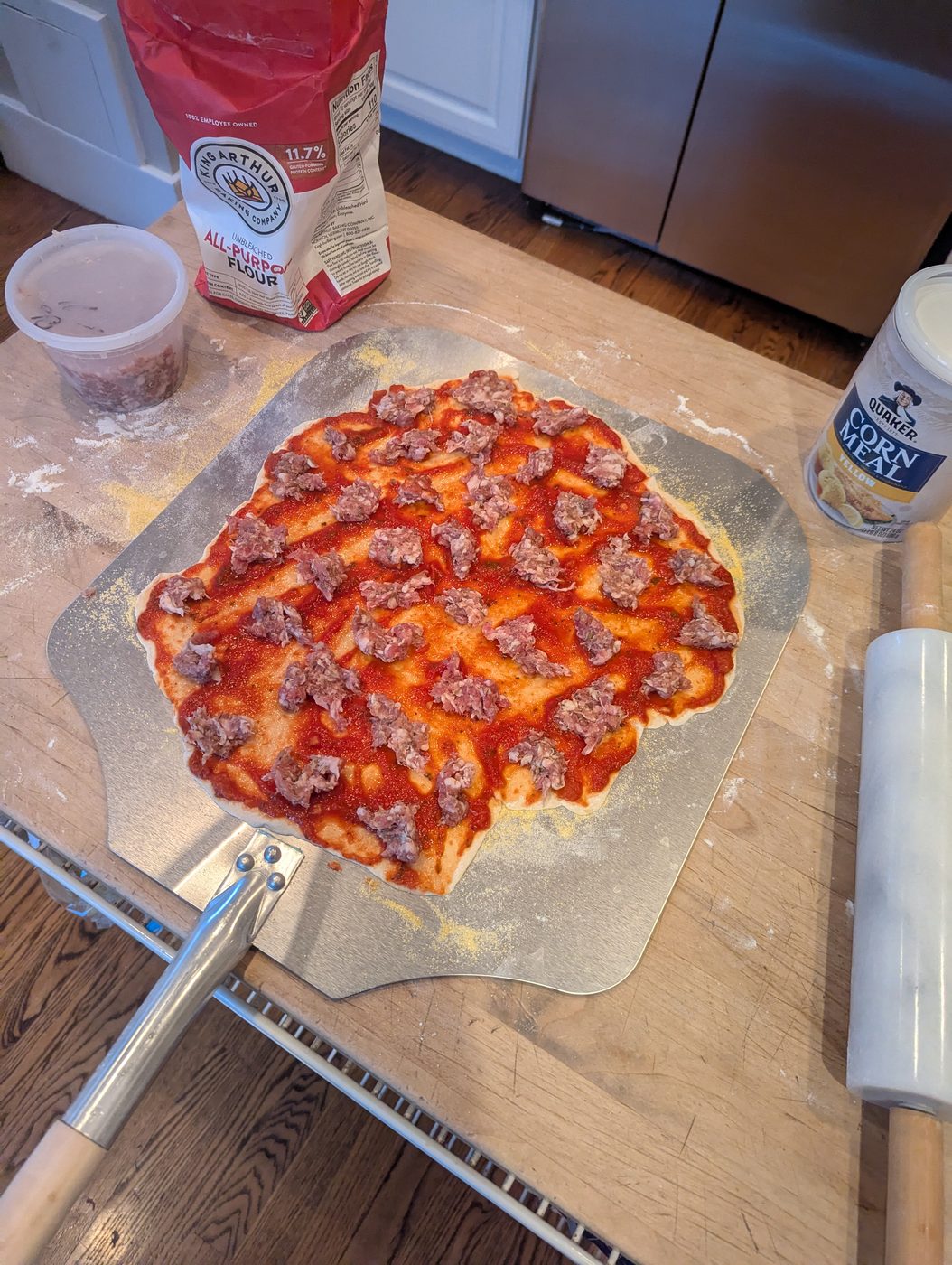 A pizza dough with sauce spread edge to edge, dense blobs of raw sausage scattered across, flour and cornmeal in the background